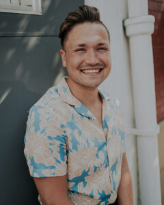 Nathan stands in front of a gray wall wearing a blue and peach floral pattern shirt. He is a white, male presenting human, smiling.