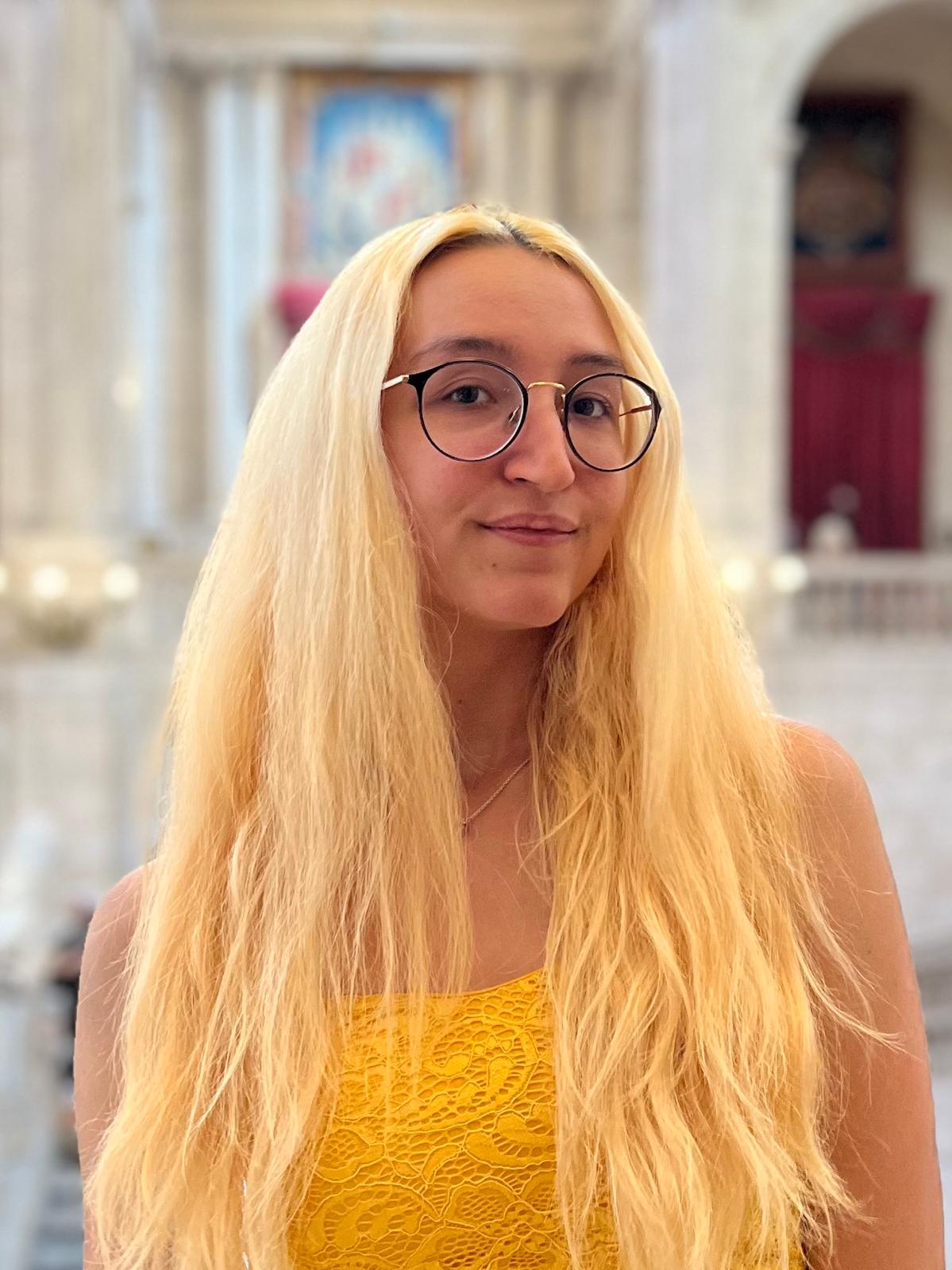 A white woman with long bright blonde hair, dark eyes, and wearing round-shaped glasses. She wears a bright-yellow top without sleeves. On the background, she has a right hall with white walls and arcs.